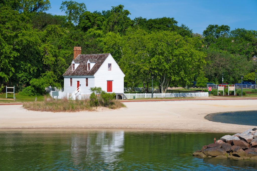The historic Archer House, a white wooden cottage with red doors and a brick chimney, stands on a sandy shoreline next to calm water, surrounded by lush green trees.