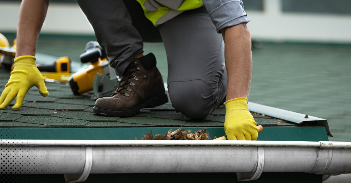 Worker cleaning debris from gutters while wearing gloves and safety gear.