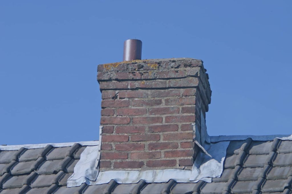 A brick chimney with visible masonry wear and a metal flue pipe sits atop a tiled roof against a clear blue sky.