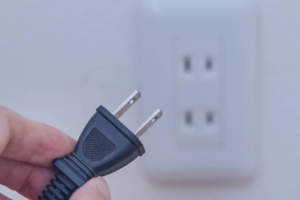 A close-up of a hand holding a black two-prong electrical plug in front of a blurred white wall outlet.